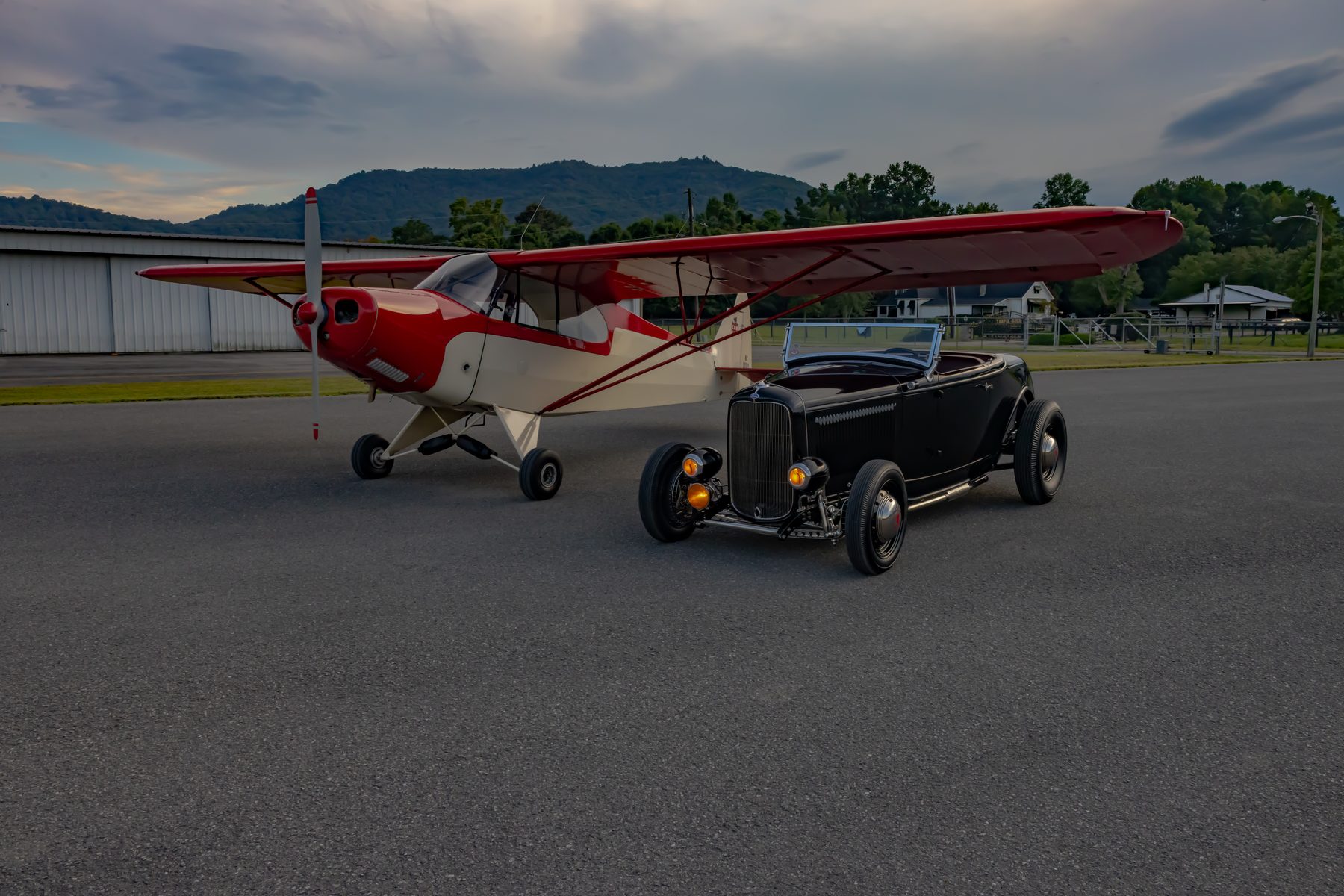 Black hot rod parked beside a vintage red and white Piper airplane